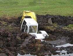 Photograph of a backhoe that is over fifty percent submerged in a large hole that it dug in a peat bog before falling in.