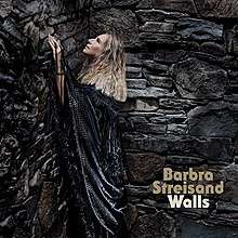 Barbra Streisand wears a black shall, gazing upward, surrounded by a large stone wall