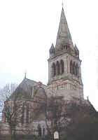 Photo of a church and tower with spire, partially hidden by the bare branches of trees.
