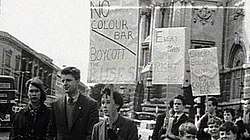 A group of young people with placards supporting boycott on the street. Behind them on the upper right is a portion of a large stone building, on the upper left the upper floors of a terrace. Behind the people, at street level, motor cars and buses can be seen.