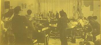 A somewhat indistinct photo on yellow paper of a crowded meeting room, showing student protesters with signs.  In the front, several well-dressed individuals appear to be gathering papers and departing.