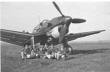 a single-engine monoplane parked on grass with men in uniform seated under the fuselage