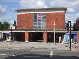 A red-bricked building with a rectangular, grey sign reading "SUDBURY HILL STATION" in black letters all und er a blue sky with white clouds