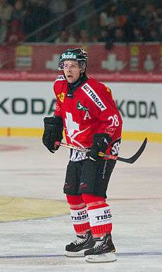 A Caucasian male ice hockey player shown skating on ice in a full-body shot. He is wearing a red and white sweater with a dark helmet.