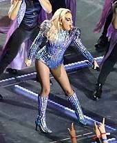 Lady Gaga in the jumpsuit pointing her microphone towards the audience from onstage.