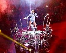 Lady Gaga standing atop a skeletal structure and singing, as red light is seen behind her.
