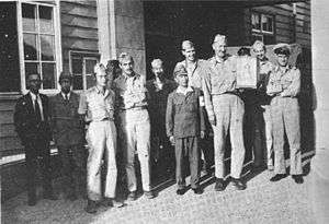 A group of men in uniforms stand in front of a wooden building. One, holding a box, is very tall, towering over the Asian man next to him.