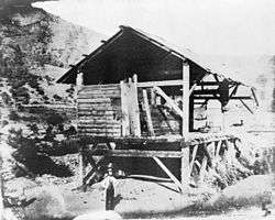 Historic photograph of Sutter's Mill in Coloma in 1850. It is a largely open-walled wooden building set on stits among sparsely vegetated mountains.