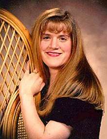 A young white woman with straight reddish-tinted blonde hair that runs down below her shoulders over a black top smiles and looks at the camera. She is turned sideways with her left shoulder in front; her left arm is crooked up to hold onto a wooden latticed object at the left side.