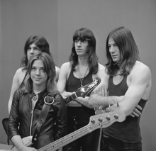 A black and white photograph of Quatro and her unnamed backing band. Quatro is holding her bass guitar, standing, and wearing a black leather jacket; her three taller and long-haired male band members are standing behind her wearing dark tee shirts.