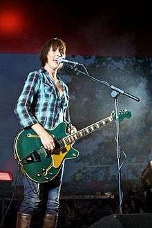 A three-quarter shot of a woman singing into her microphone, she holds her right had over her guitar strings, her left hand is obscured behind the guitar at her hip. She wears a blue-black check shirt and jeans, which are tucked into her brown boots. Her microphone stand is at her left. The background is darkened, with barely visible trees and people, some lighting and equipment is visible.