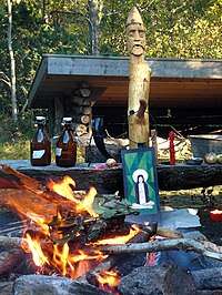 An outdoor fire burning in front of a wooden post with an anthropomorphic face carved into the top