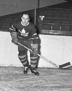 A man in full hockey gear skates toward the camera.  He is in a dark jersey with a stylized maple leaf logo and is holding his stick left handed.