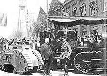 A military man in full dress uniform salutes a suited business man who is doffing his bowler hat in return. Behind the two men is a Holt tractor, which towers above them, and a diminutive one-man tank, which is below chest height. Behind the vehicles, a crowd looks on.
