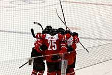 A group of hockey players stand on the ice in a circle, hugging each other and celebrating. They are all wearing red and white sweaters.