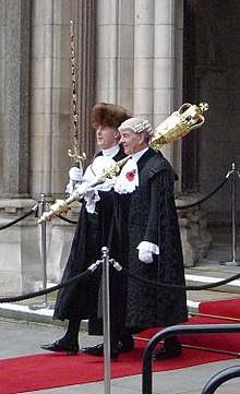 Two men in black cloaks walking side by side on a red carpet. One wears a fur hat and carries a sword in a red and gold scabbard upright. The other wears a judge's wig and has a large, gold mace over his left shoulder.