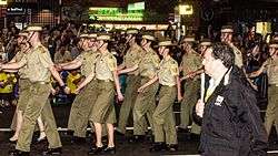 Colour photo of men and women marching down a street while wearing green military uniforms