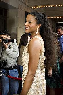 A woman wearing a tan/gold dress. She is looking toward the left, while smiling toward cameras.