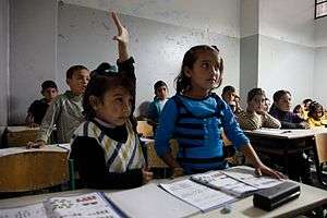 Children at desks in a classroom.  One child raises her hand.