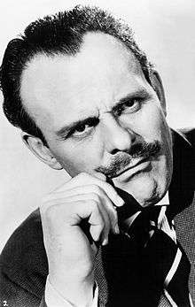 Terry-Thomas, wearing a dark jacket, looks off to the right, cuffing his moustache