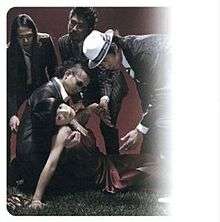 A woman collapsed on the ground in a formal dress, looking at the camera. Four men in suits are attending to her.