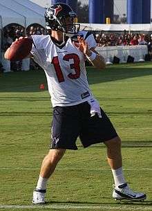 An American football player throwing a football. He is wearing a white jersey with the number 13 across the chest.