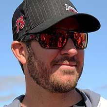 A bearded man in his mid thirties wearing black sunglasses and a black baseball cap with the number 78 on his right-hand side
