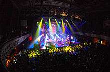 Multiple colored lights shine down on a stage while an audience watches.