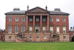 A large Palladian-style house with three storeys, the bottom storey in stone, the upper storeys in brick, with a prominent portico with columns rising to the full height of the building