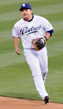 A man wearing a white baseball uniform with "Padres" in navy blue script across the chest and a blue baseball cap atop his head