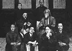 Posed group black-and-white photograph of seven Chinese men, possibly academics, in formal wear: two wear European-style suits, the five others wear Chinese traditional dress; four of the seven sit on the floor in the foreground; another sits on a chair behind them at centre-left; two others stand in the background. They surround an eighth man who is robed, bearded, and sitting in a chair placed at centre-left. Four elegant windows are behind them in a line.