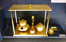 A photograph of several golden vessels, drinking cups, a guilted ladle and instruments, are displayed on a stand. Beside the stand is a golden plate and cup on a golden saucer