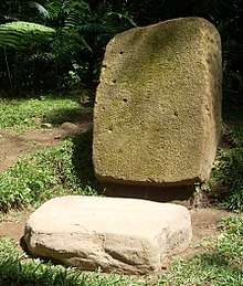 A moss-covered upright stone slab, fallen slightly from vertical. The slab has a number of small cup-like depressions in the surface. In front of it is a horizontal roughly square slab of pale stone with faint designs sculpted around the edges.