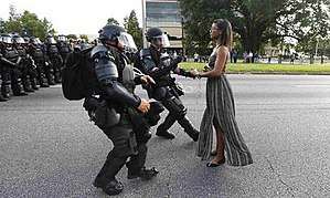 Ieshia Evans in a flowing dress stands facing a line of police in riot gear, two of whom charge towards her