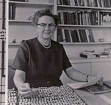 A woman with glasses sits at a table with a tray of specimen vials. A bookshelf is behind her.