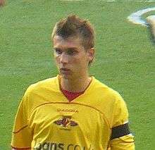 The head and upper body of young man wearing a yellow top, standing on a grass field. His top features a distinctive red, black and yellow crest, with the word "Watford" visible below it.