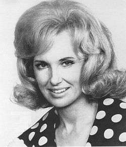 A blonde woman wearing a dark blouse with polka dots, smiling slightly