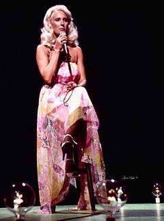 A woman with long blonde hair, wearing a long multi-coloured dress, sitting on a stool and singing into a microphone
