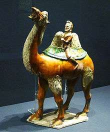 A glazed figurine of a red camel, which is being ridden by a bearded merchant in green clothing