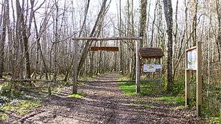 photo showing a path going straight ahead through a wood, with a car-wide portal across it made of wooden posts and from whose transversal pole hangs on chains a wooden sign that bears the inscription: "Le ferrier". On the right hand-side of the portal is a small tile-roofed shelter for a sign that indicated the various major parts of the ferrier. On the left hand-side of the portal is a gap to let walkers, horses and pushbike-riders through. On the left of this gap is a low barrier, also made of wooden poles, about 2 metre wide. Some 2 meters before the portal on the right hand-side of the path is another sign that stands parallel to the road; the photo does not show what that sign displays (it is a map of the orienteering tracks and gives basic indications about the sport as practiced on this site)
