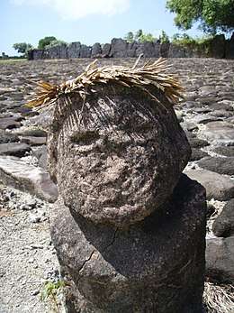 Weathered grey stone bust with a palm frond crown.  Grey rocks with white and in between them in the background.