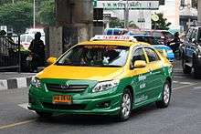 A Toyota saloon/sedan driving on the road, its top half painted in yellow and the bottom half in green, with a sign on the roof saying &quot;TAXI-METER&quot; and a yellow licence plate