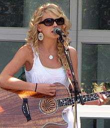 A young blonde-haired woman wearing dark glasses and a white tank top, playing a guitar and singing into a microphone