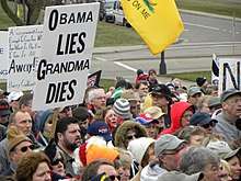 A group of people near a street corner in the background, seen from above head level. At the left one holds up a black-on-white sign saying "Obama Lies, Grandma Dies". Portions of other signs are visible and part of a Gadsden flag hangs from above the top of the image