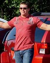 A dark haired young man wearing dark sunglasses poses for cameras with his arms outstretched. He is wearing a red T-shirt, with a pattern on both shoulders and the top part of the chest, and jeans.