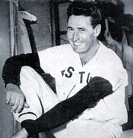 A man, wearing a white baseball uniform with the words "BOSTON" across his chest obscured, smiles towards the left.