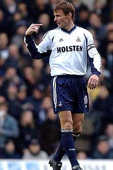 Teddy Sheringham – wearing a long-sleeved white jersey, dark blue shorts and a Premier League captain's armband on the left arm – points at himself.