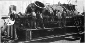 A large piece of machinery is in operation. Six workers can be seen attending to the machine while a ladder lies against it.