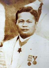 Middle aged Filipino American male wearing a white suite with a neck order medal, and two other medals on the lapel.