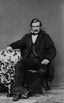 Mustached man in three-piece suit, seated.